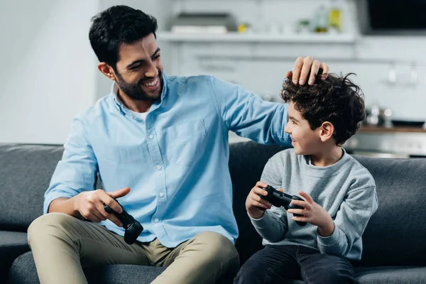 Feliz pai latino tocando o cabelo do filho bonito e segurando joysticks em casa — Fotografia de Stock