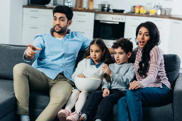 Homem latino apontando com o dedo enquanto assiste tv com a família hispânica em casa — Fotografia de Stock