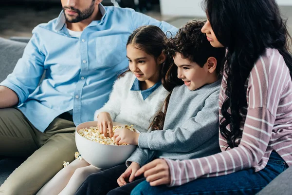Веселая латиноамериканская семья смотрит телевизор с чашей попкорна дома — стоковое фото