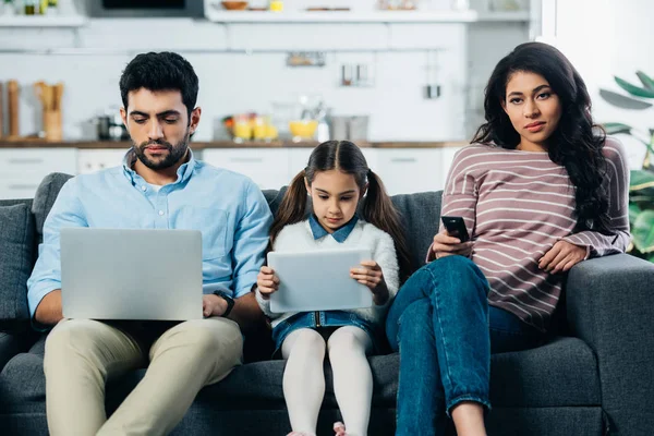 Mujer latina sosteniendo el control remoto mientras está sentada en el sofá cerca del marido con el ordenador portátil y la hija con la tableta digital - foto de stock