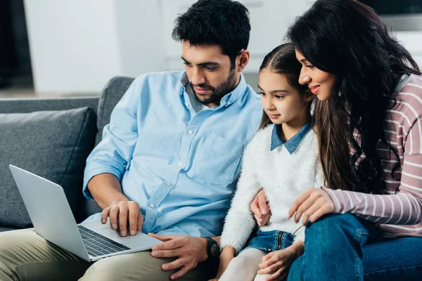 Donna latina seduta sul divano con figlia vicino al marito utilizzando il computer portatile — Foto stock