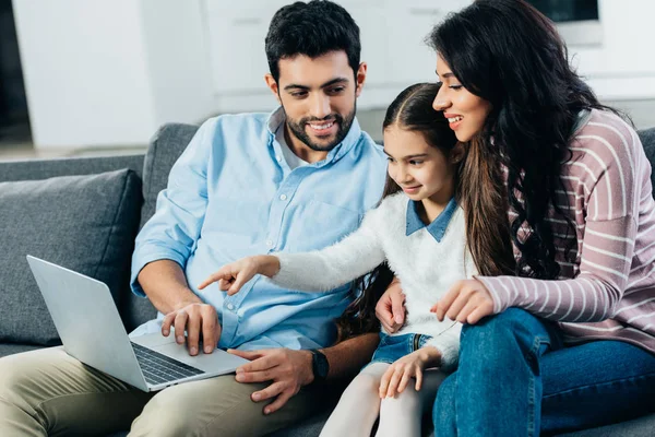 Latin woman sitting on sofa with daughter pointing with finger at laptop near dad — Stock Photo