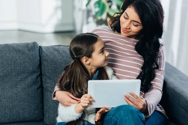 Latin woman sitting on sofa and looking at cute daughter while holding digital tablet at home — Stock Photo