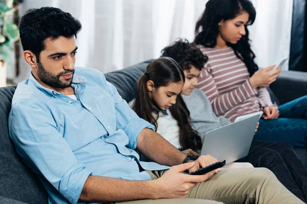 Enfoque selectivo del hombre latino guapo sosteniendo control remoto cerca de martillo hispano usando gadgets - foto de stock