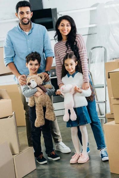 Happy latin kids holding soft toys and standing with cheerful mom and dad in new home — Stock Photo