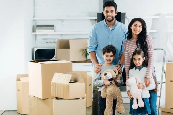 Niños latinos alegres sosteniendo juguetes suaves y de pie con mamá y papá felices en un nuevo hogar - foto de stock