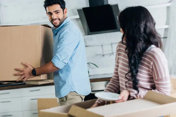 Selective focus of handsome latin man holding box near wife in new home — Stock Photo