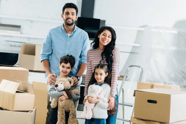 Cheerful latin children holding soft toys and standing with happy mom and dad in new home — Stock Photo