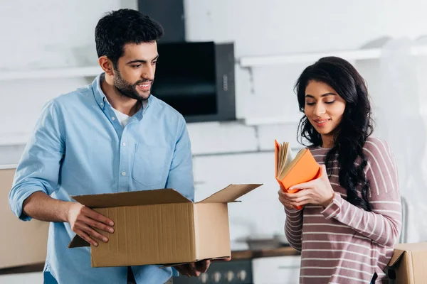 Attractive latin woman holding book while standing near husband with box in new home — Stock Photo