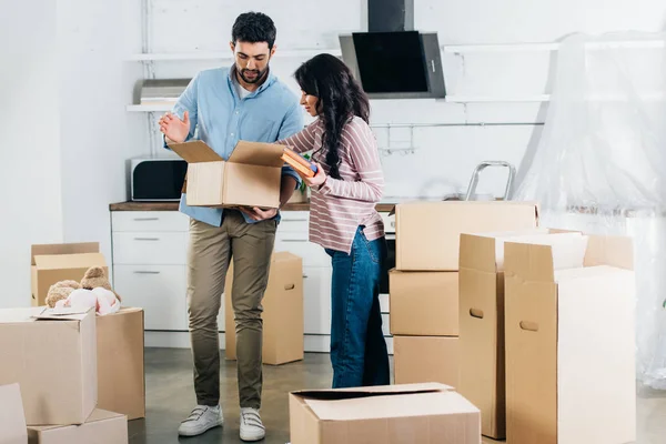 Lateinische Frau hält Bücher, während sie neben Ehemann mit Schachtel im neuen Zuhause steht — Stockfoto