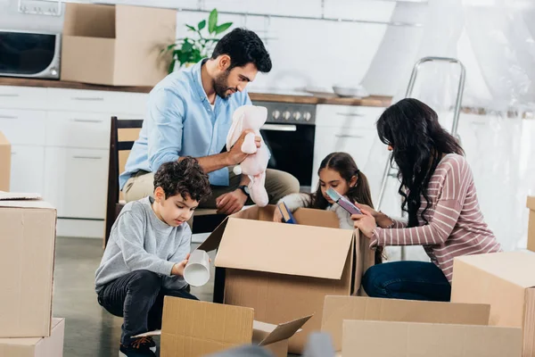 Счастливый латинский отец держит мягкую игрушку рядом с испаноязычной семьей, распаковывая коробки в новом доме — стоковое фото