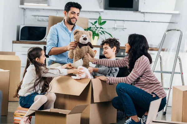 Alegre pai latino segurando brinquedo macio perto da família hispânica enquanto desembalando caixas em nova casa — Fotografia de Stock