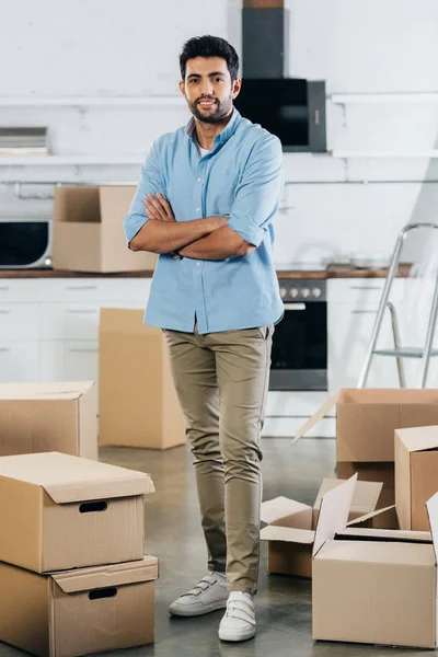 Cheerful latin man standing with crossed arms near boxes while moving in new home — Stock Photo