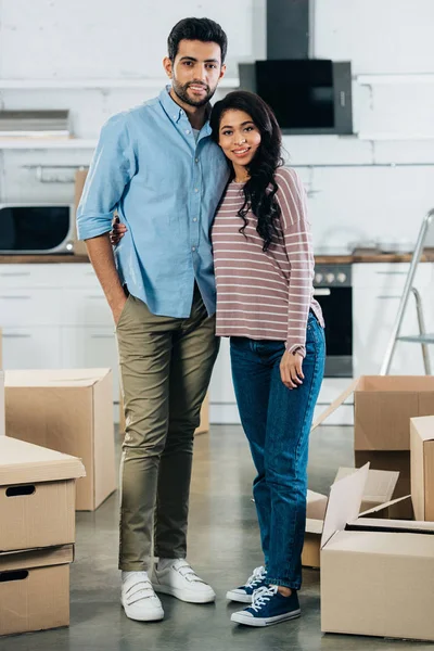 Cheerful latin couple standing and smiling near boxes in new home — Stock Photo