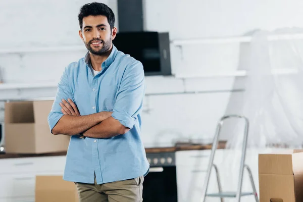 Handsome latin man standing with crossed arms near boxes while moving in new home — Stock Photo