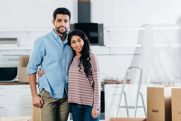 Cheerful latin couple embracing and smiling near boxes in new home — Stock Photo