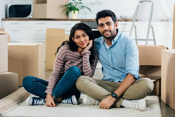 Alegre pareja latina sentada con las piernas cruzadas cerca de cajas en nuevo hogar - foto de stock