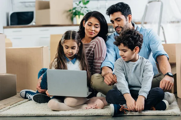 Cheerful latino family looking at laptop while sitting on carpet in new home — Stock Photo