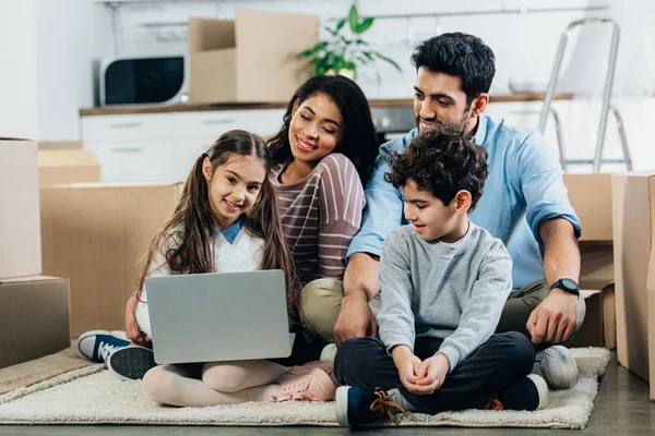 Alegre familia hispana mirando a la computadora portátil mientras está sentado en la alfombra en un nuevo hogar - foto de stock