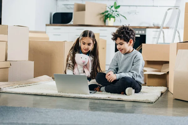 Niños felices usando el ordenador portátil mientras están sentados en la alfombra en un nuevo hogar - foto de stock