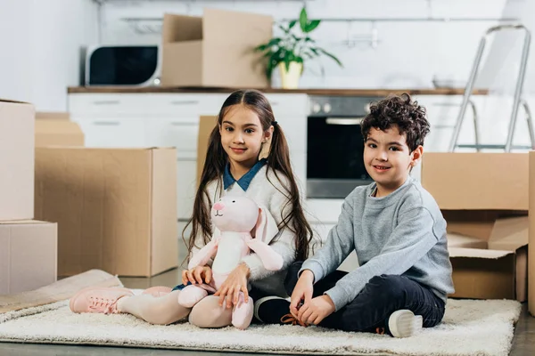 Crianças felizes sentadas no tapete com brinquedo macio perto de caixas enquanto se movem em nova casa — Fotografia de Stock