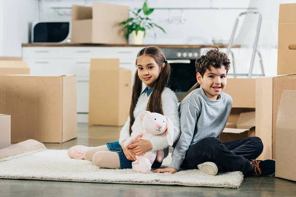 Happy kids sitting on carpet with soft toy near boxes while moving in new home — Stock Photo