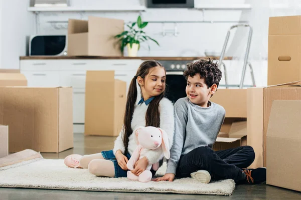 Happy kids sitting on carpet with soft toy and looking at each other in new home — Stock Photo