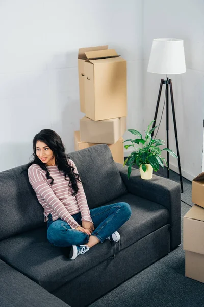 Cheerful latin woman sitting on sofa with crossed legs in new home — Stock Photo