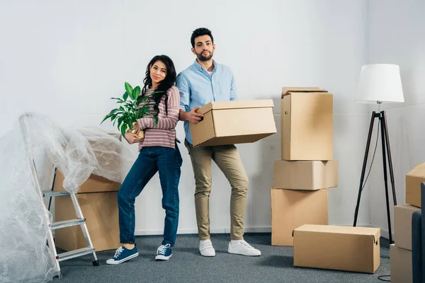 Happy latin woman holding plant near husband with box while moving into new home — Stock Photo