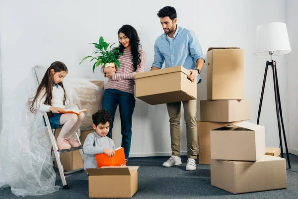 Cheerful latin mom and dad looking at cute kids reading books in new home — Stock Photo