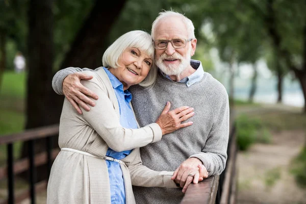 Sourire couple aîné étreignant tout en se tenant près de rambarde pont en bois et en regardant la caméra — Photo de stock