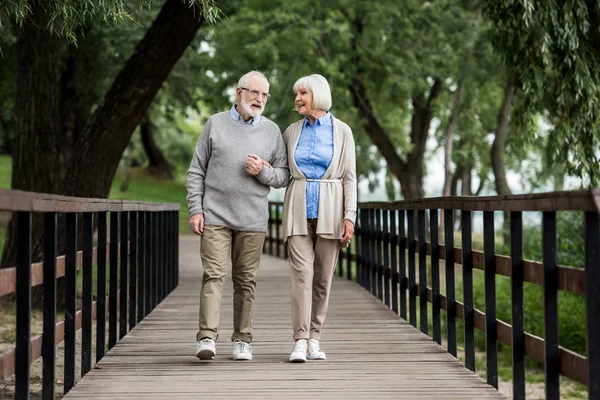 Улыбающаяся пожилая пара разговаривает во время прогулки по деревянному мосту в парке — стоковое фото