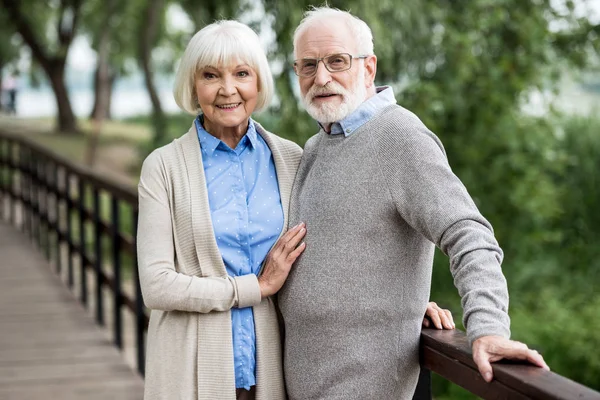 Selective focus of nice smiling senior couple standing on wooden bridge in park — Stock Photo