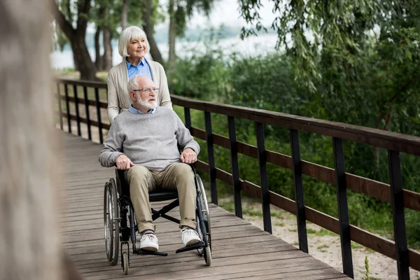Mujer mayor sonriente con marido en silla de ruedas mientras camina en el parque - foto de stock