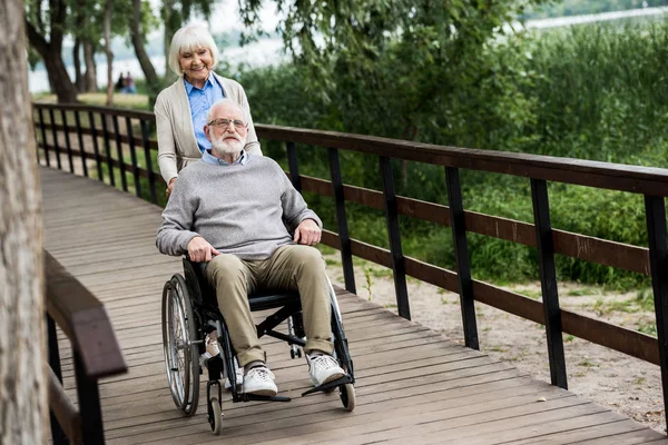 Donna anziana sorridente con marito in sedia a rotelle sul ponte di legno nel parco — Foto stock