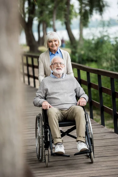 Selektiver Fokus einer netten Seniorin mit Mann im Rollstuhl auf Holzbrücke — Stockfoto