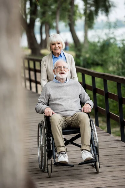 Selective focus of nice senior woman with husband in wheelchair on wooden bridge — Stock Photo