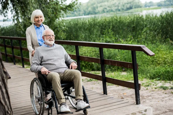 Happy senior man in wheelchair with smiling wife on wooden bridge in park — Stock Photo