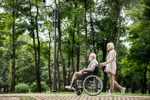 Mujer mayor llevando marido en silla de ruedas mientras camina en el parque - foto de stock