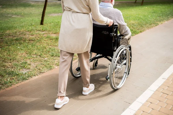 Vista recortada de la mujer mayor llevando marido en silla de ruedas mientras camina en el parque - foto de stock