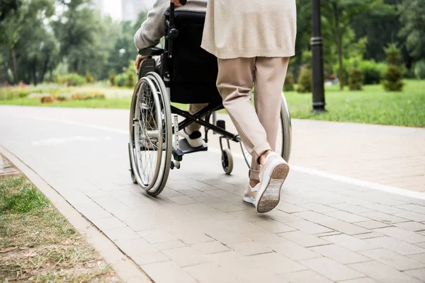 Vista parcial de la mujer mayor que lleva al marido en silla de ruedas mientras camina en el parque - foto de stock