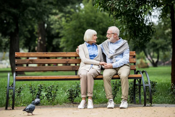 Senior couple talking and smiling while sitting on wooden bench in park — Stock Photo
