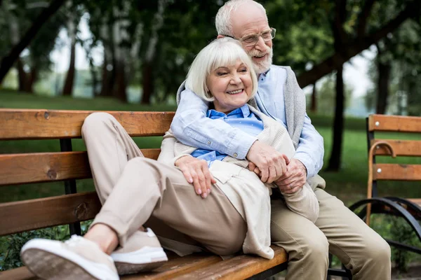 Sonriente mujer mayor acostado en banco de madera cerca feliz marido en parque - foto de stock