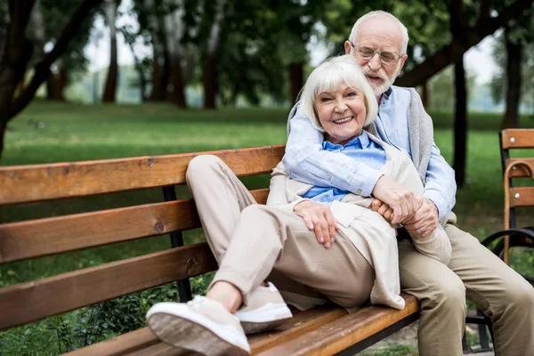 Happy senior woman laying on wooden bunch near smiling husband in park — Stock Photo