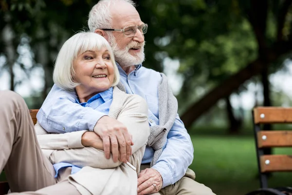 Homme âgé souriant embrassant femme heureuse tout en étant assis ensemble sur le banc dans le parc — Photo de stock