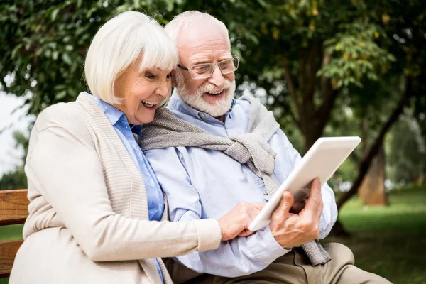 Улыбающаяся пожилая пара с помощью цифрового планшета, сидя на скамейке в парке — стоковое фото