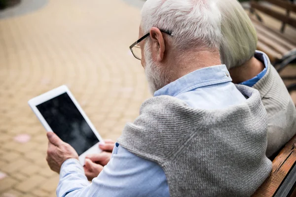 Pareja de ancianos usando tableta digital juntos mientras están sentados en el banco en el parque - foto de stock