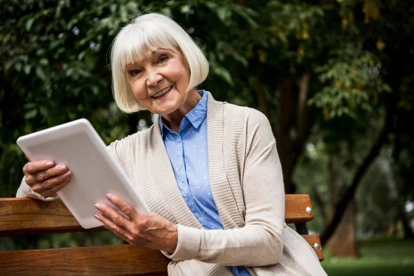 Щаслива старша жінка, використовуючи цифровий планшет, сидячи на дерев'яній лавці — стокове фото