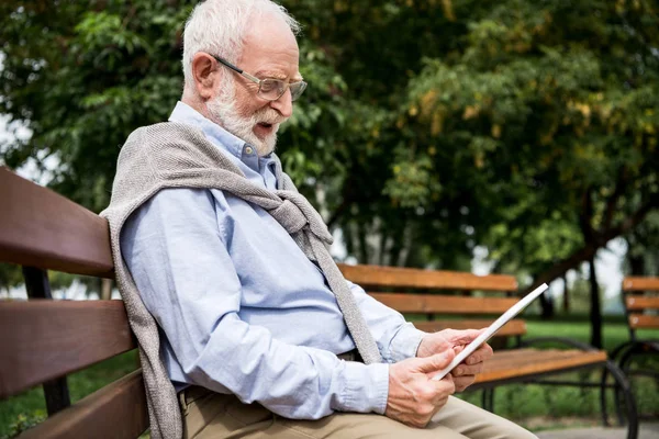 Selective focus of senior man using digital tablet while sitting on wooden bench in park — Stock Photo