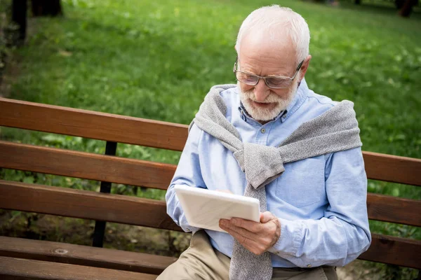 Uomo anziano sorridente utilizzando tablet digitale mentre seduto su una panchina di legno — Foto stock